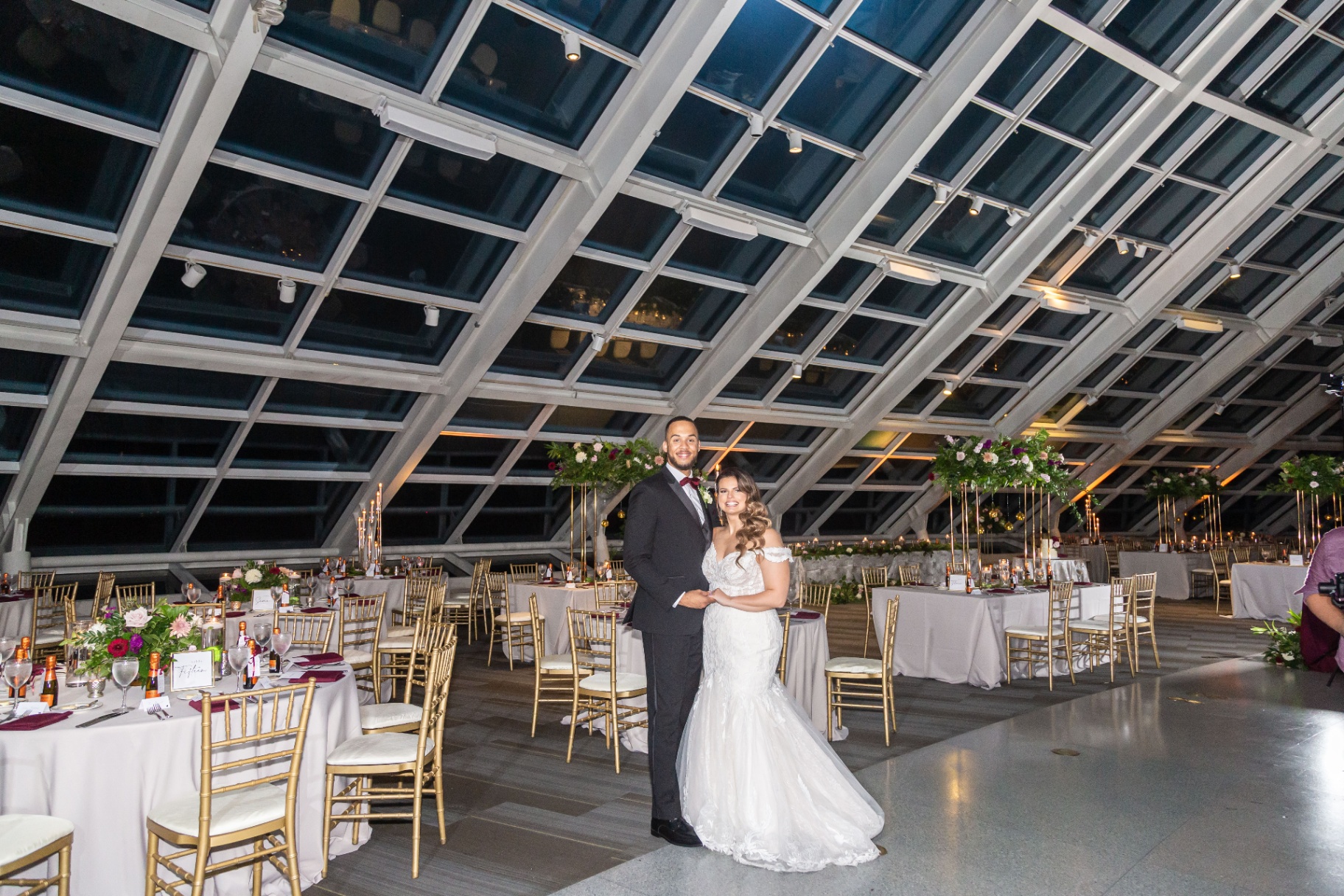 Bride and groom standing in an elegant reception room beneath a dramatic glass ceiling.